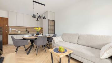 A modern living room with a grey sofa, a dining table with chairs, and a kitchen area with cabinets and a wooden countertop.