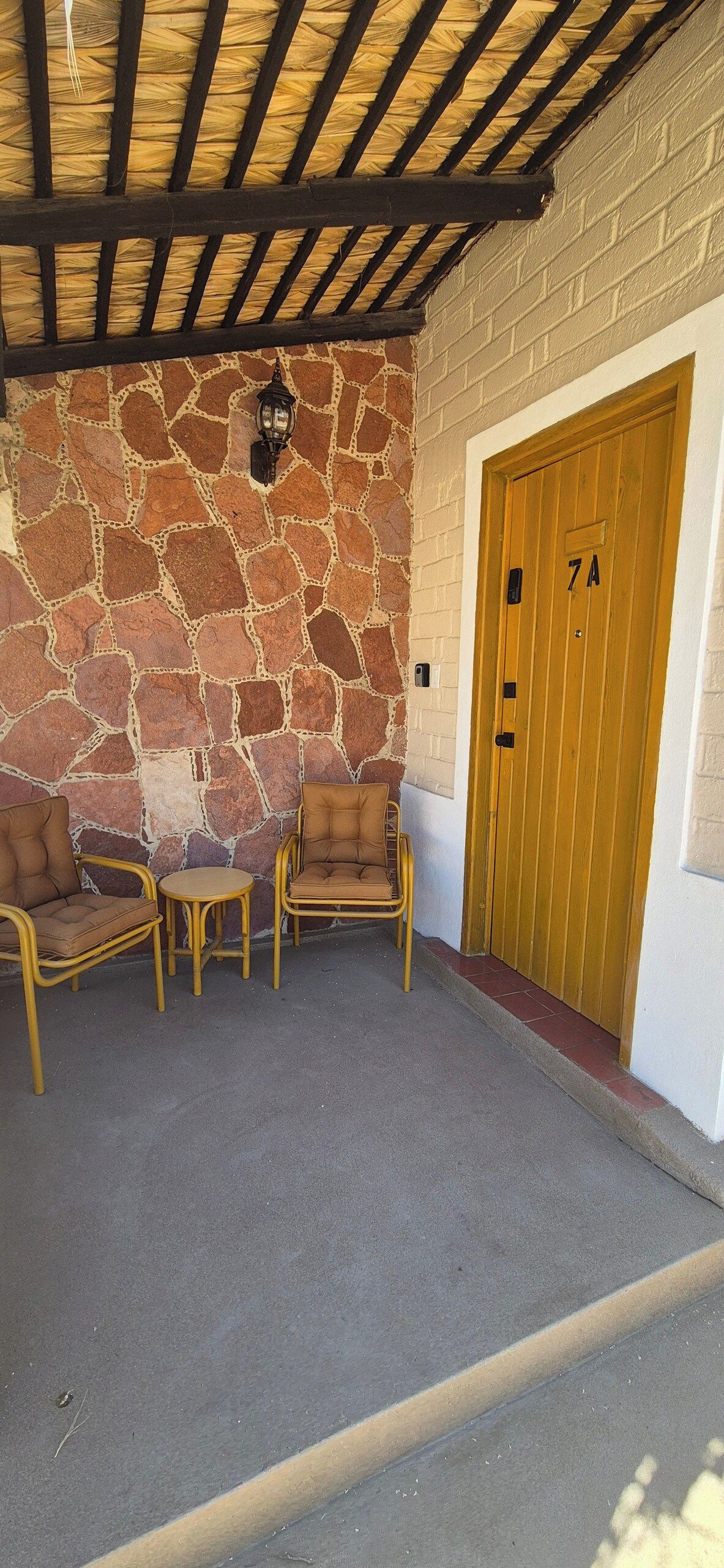 Un área de estar al aire libre techada con paredes de piedra, una puerta de madera con un número y una silla de mimbre.