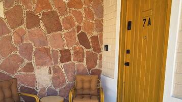 Un área de estar al aire libre techada con paredes de piedra, una puerta de madera con un número y una silla de mimbre.