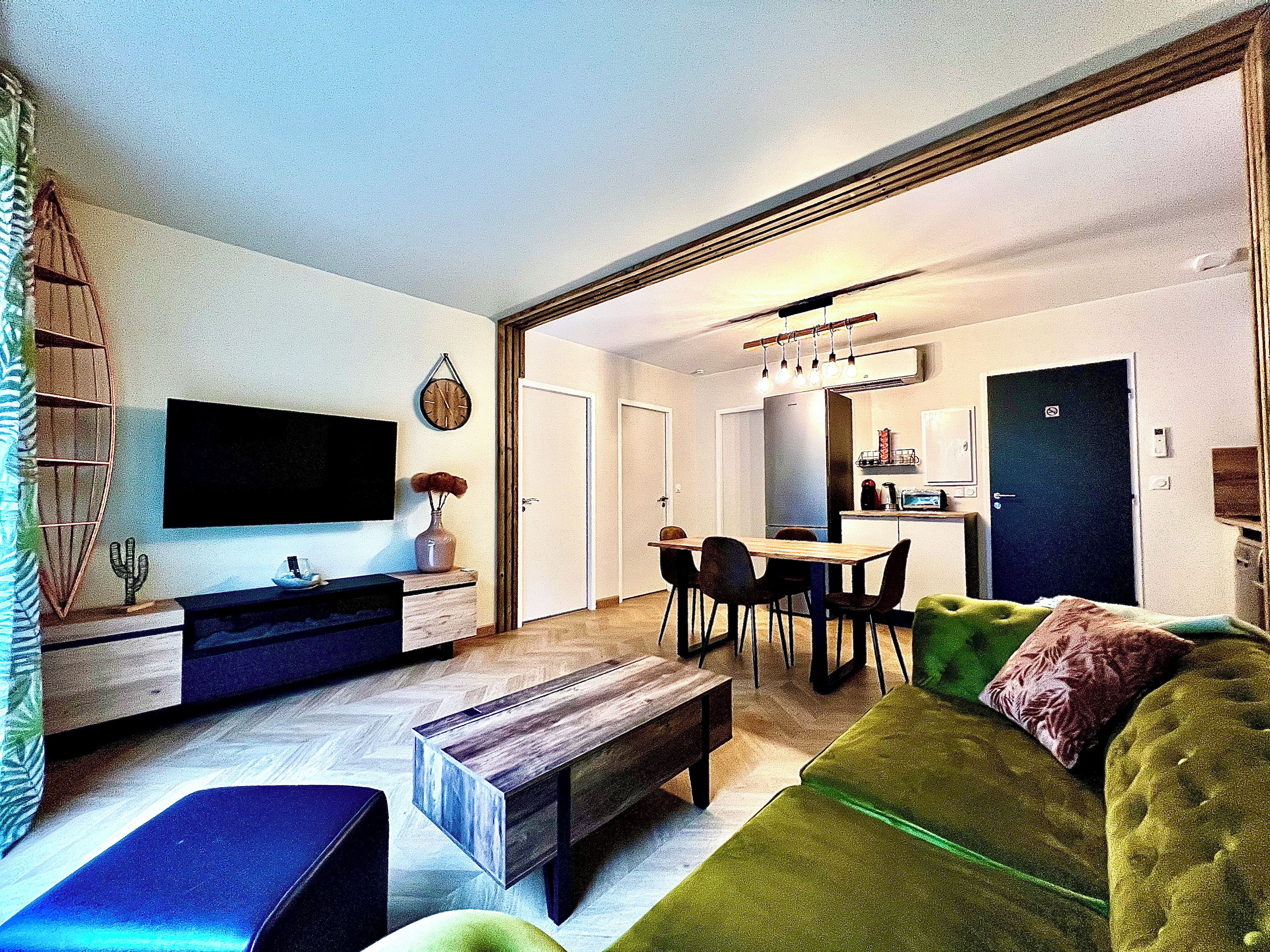 A modern living room with a flat-screen TV, a dining area with a wooden table and chairs, and a kitchen in the background.