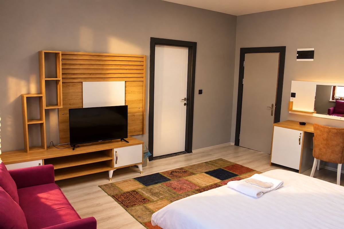 Une chambre d’hôtel moderne avec un téléviseur à écran plat posé sur un meuble en bois, un lit recouvert d’une couette blanche, un bureau avec une chaise et un tapis coloré.