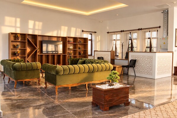 A spacious lobby with two green upholstered sofas, a wooden coffee table, and a reception desk with patterned decor.