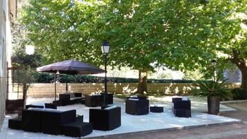 An outdoor seating area with black sofas and a large umbrella, surrounded by trees and a brick wall.