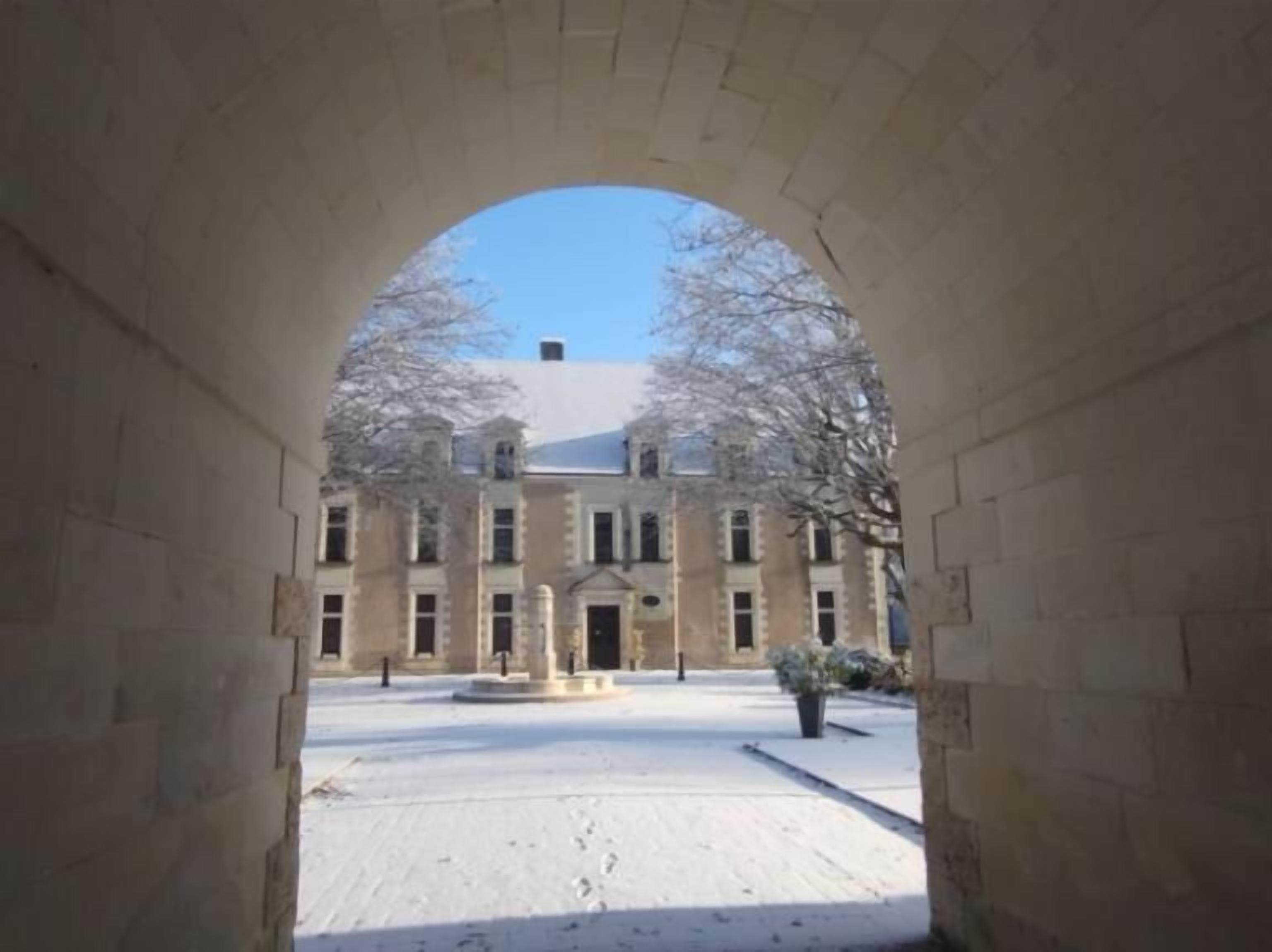 A view through an arched stone structure towards a building with a symmetrical facade, a central entrance, and a paved courtyard.