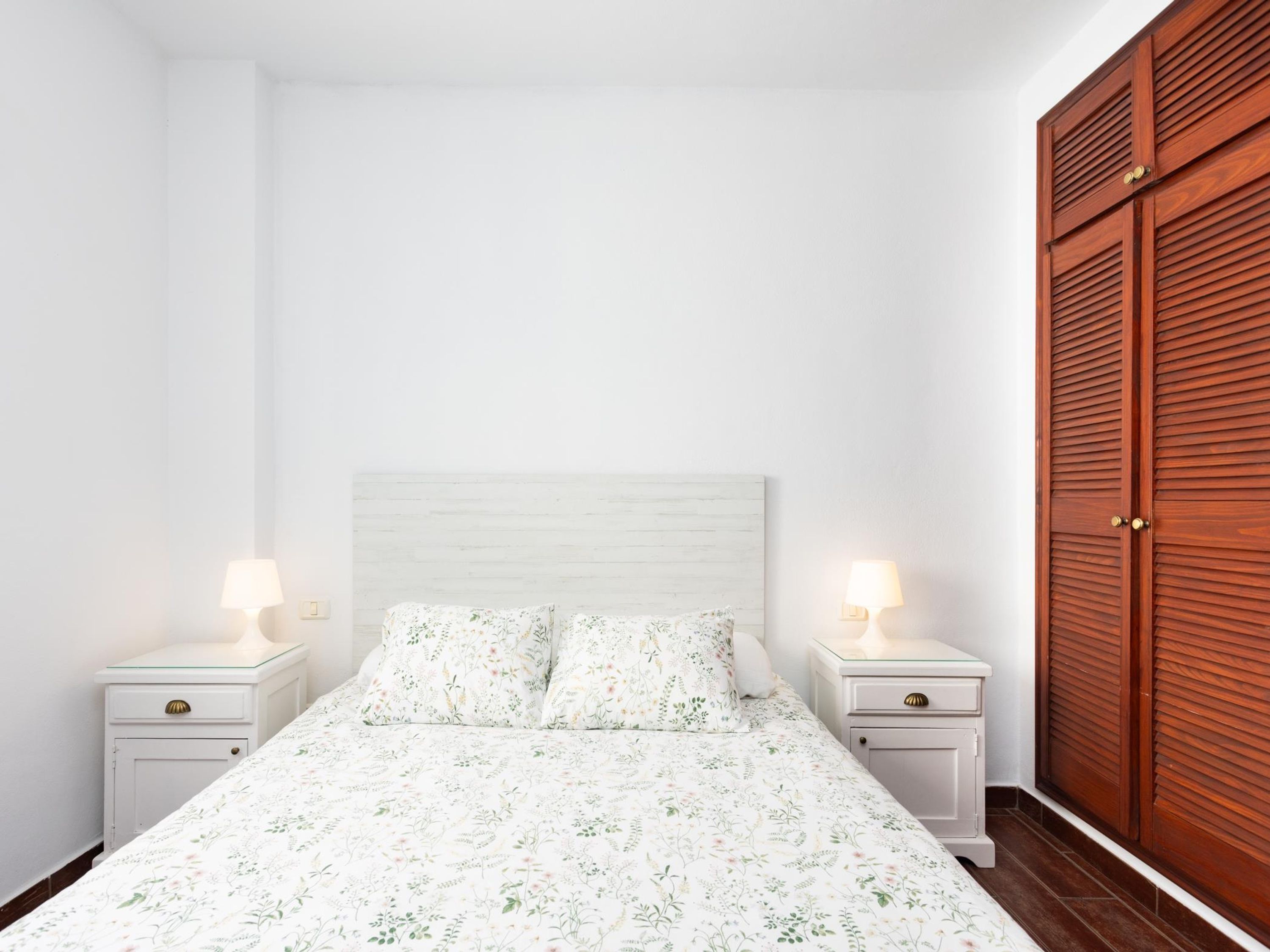 A bedroom with a bed, two bedside tables with lamps, and a wooden wardrobe.