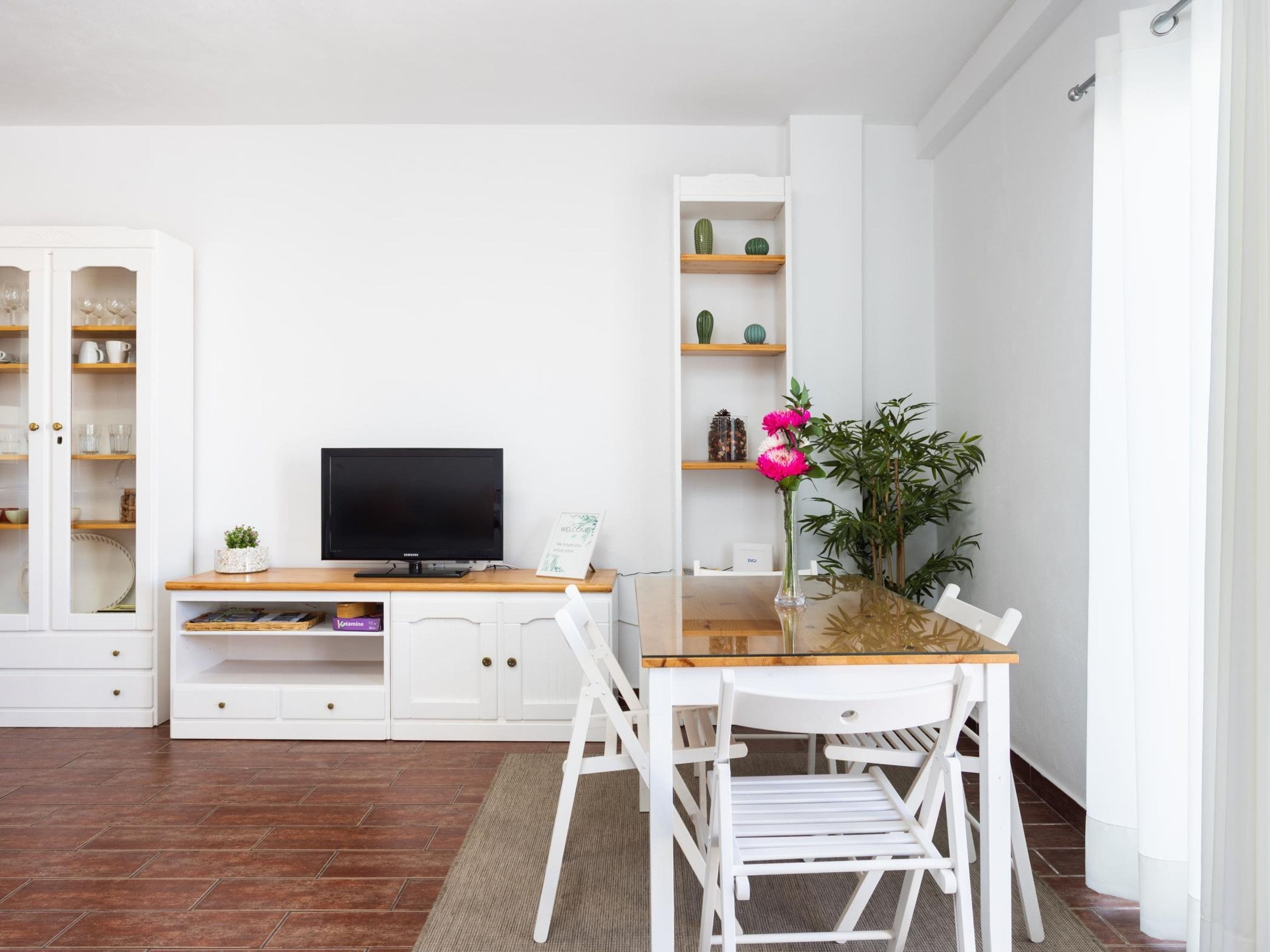 A modern living room with a flat-screen TV, white cabinetry, a dining table with chairs, and a potted plant.