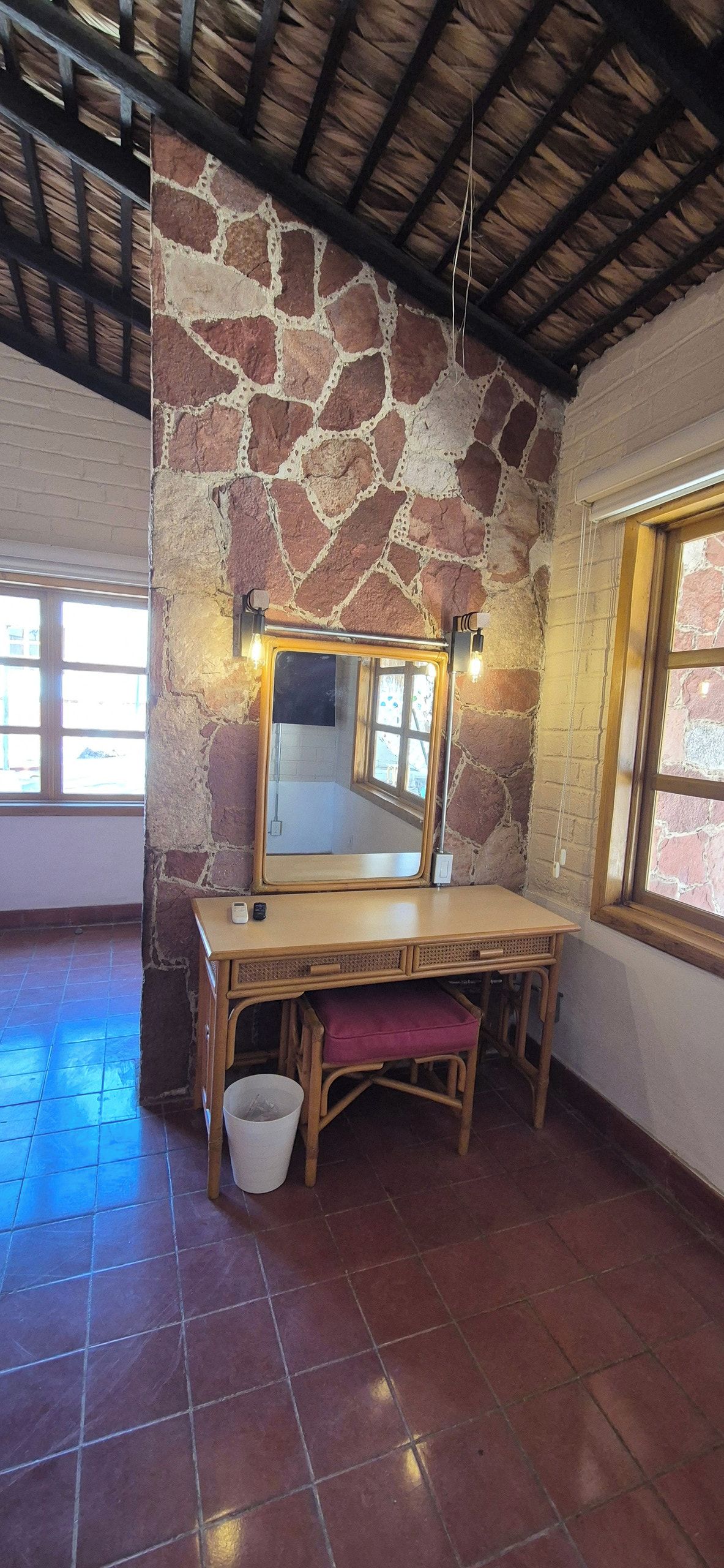 Une pièce avec un mur en pierre, un bureau en bois, un miroir, un banc et une fenêtre.