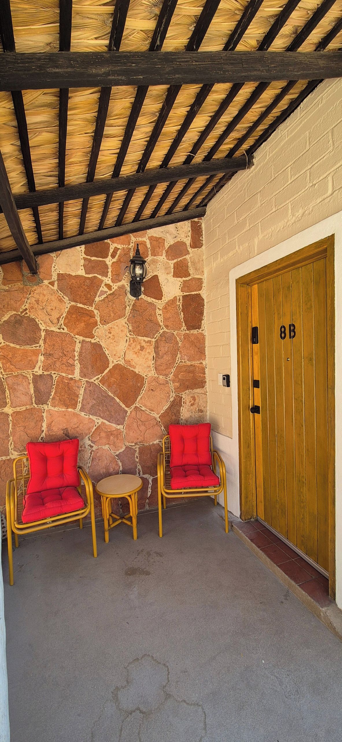 Un espace extérieur aménagé avec des sièges, abrité par un toit, doté de murs en pierre, de deux fauteuils en osier avec des coussins rouges, d’une petite table ronde et d’une porte en bois portant l’inscription « 8B ».