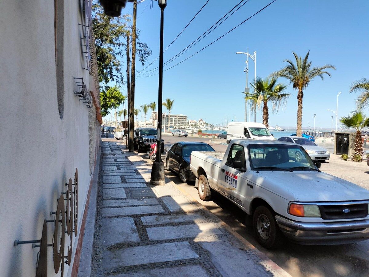 A street with parked vehicles, including a white pickup truck with 'FESACO' on the side, and a white van. There are palm trees and a clear sky.