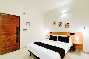 A hotel room with a wooden door, a bed with white and black bedding, two bedside tables with lamps, and three framed artworks on the wall.