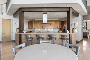 A modern kitchen with wooden cabinets, a central island with bar stools, and a dining table in the foreground. - Mountain & City Views | Hot Tub + Sauna | Trails to Mt Sanitas | Castle Black (Boulder)