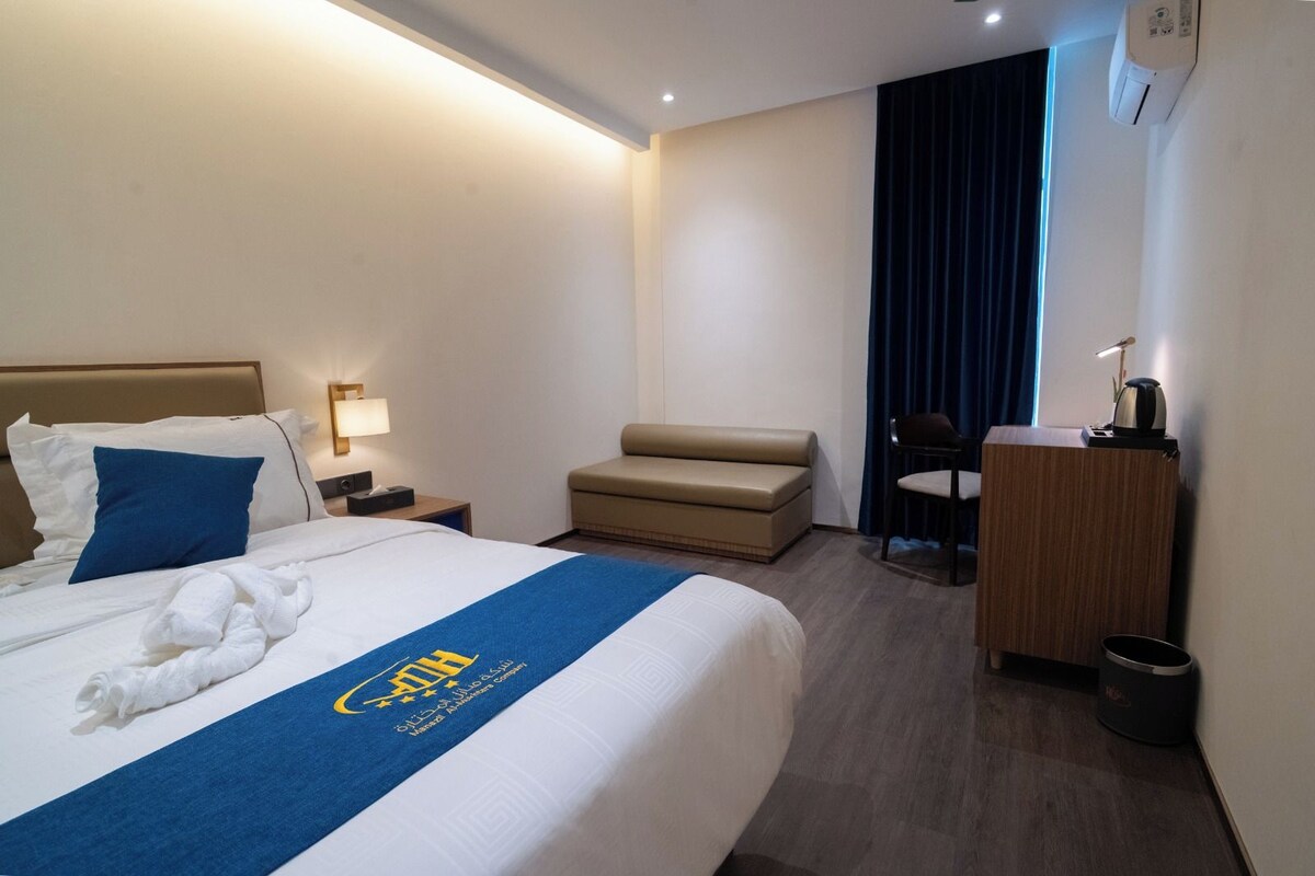Ett hotellrum med en säng, ett skrivbord med en stol, en liten soffa och ett fönster med gardiner.