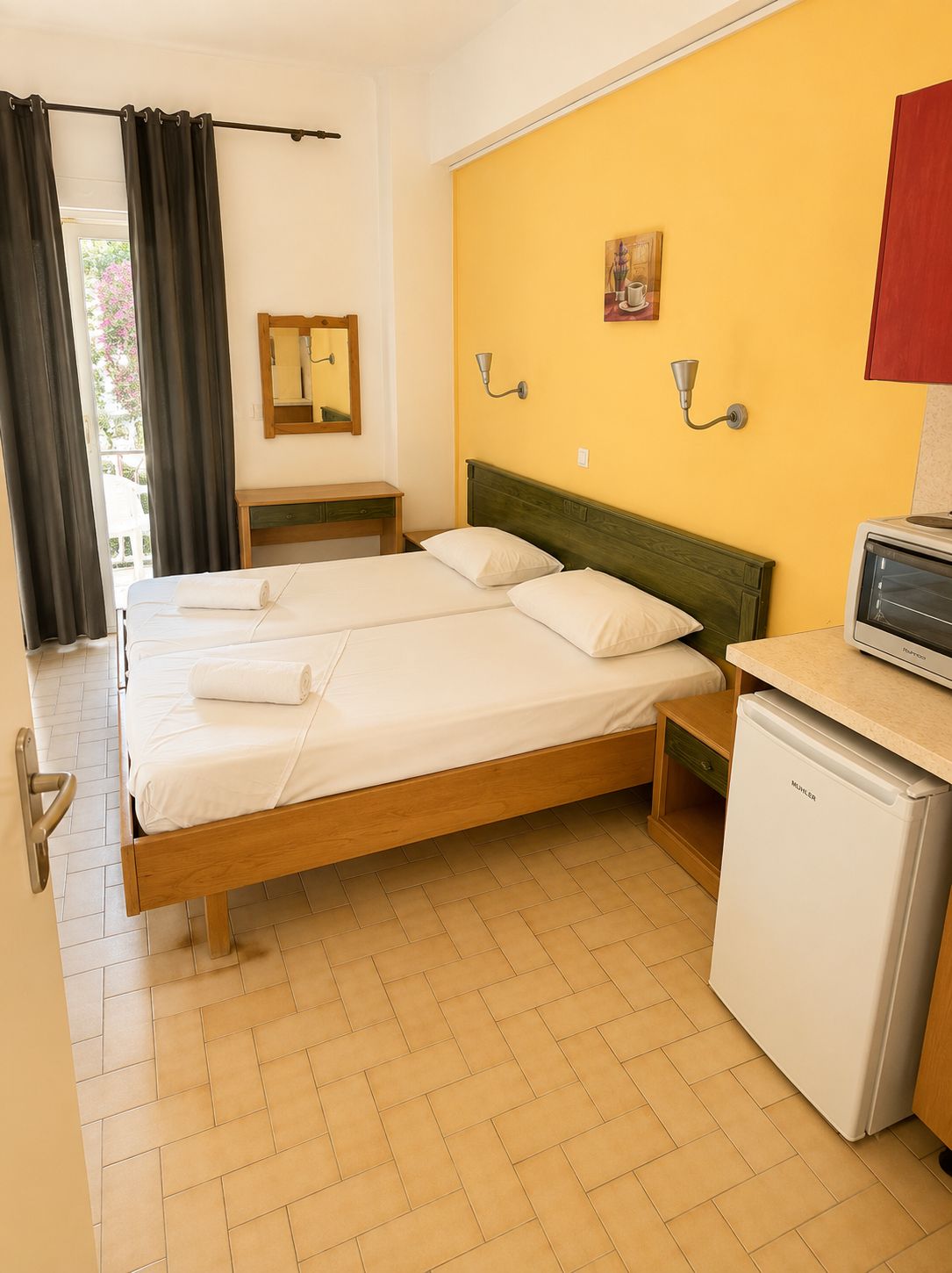 Habitación de hotel con cama doble, una mesita de noche, una lámpara de pared, un cuadro, un armario rojo y un refrigerador blanco.