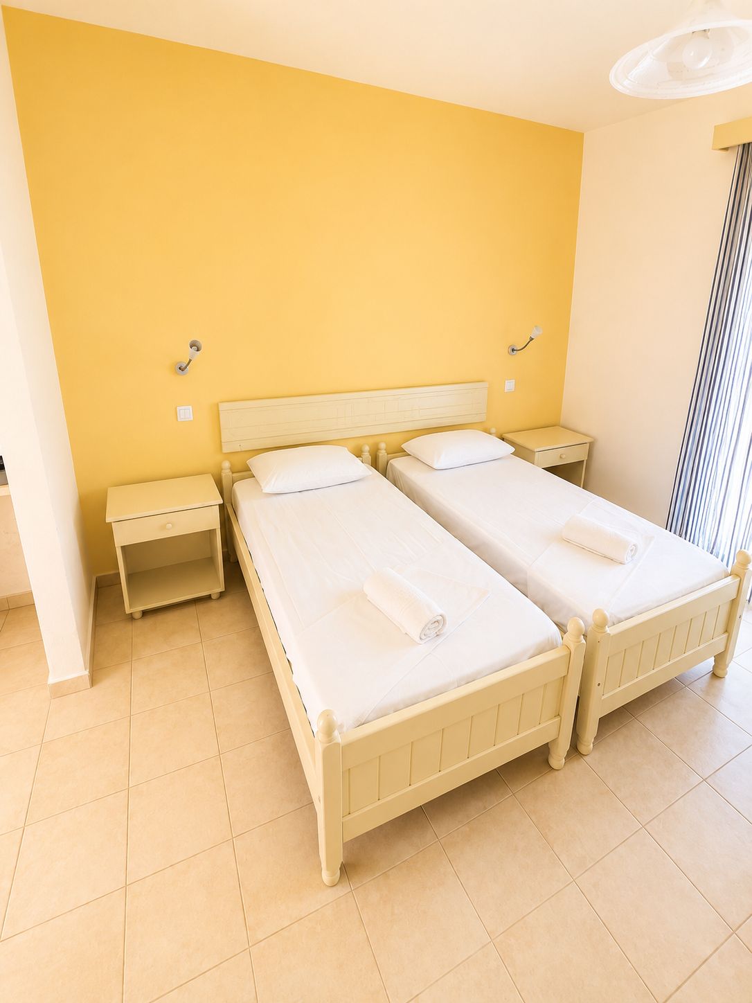A double bed with white bedding and a rolled towel, two bedside tables, and a plain yellow wall.