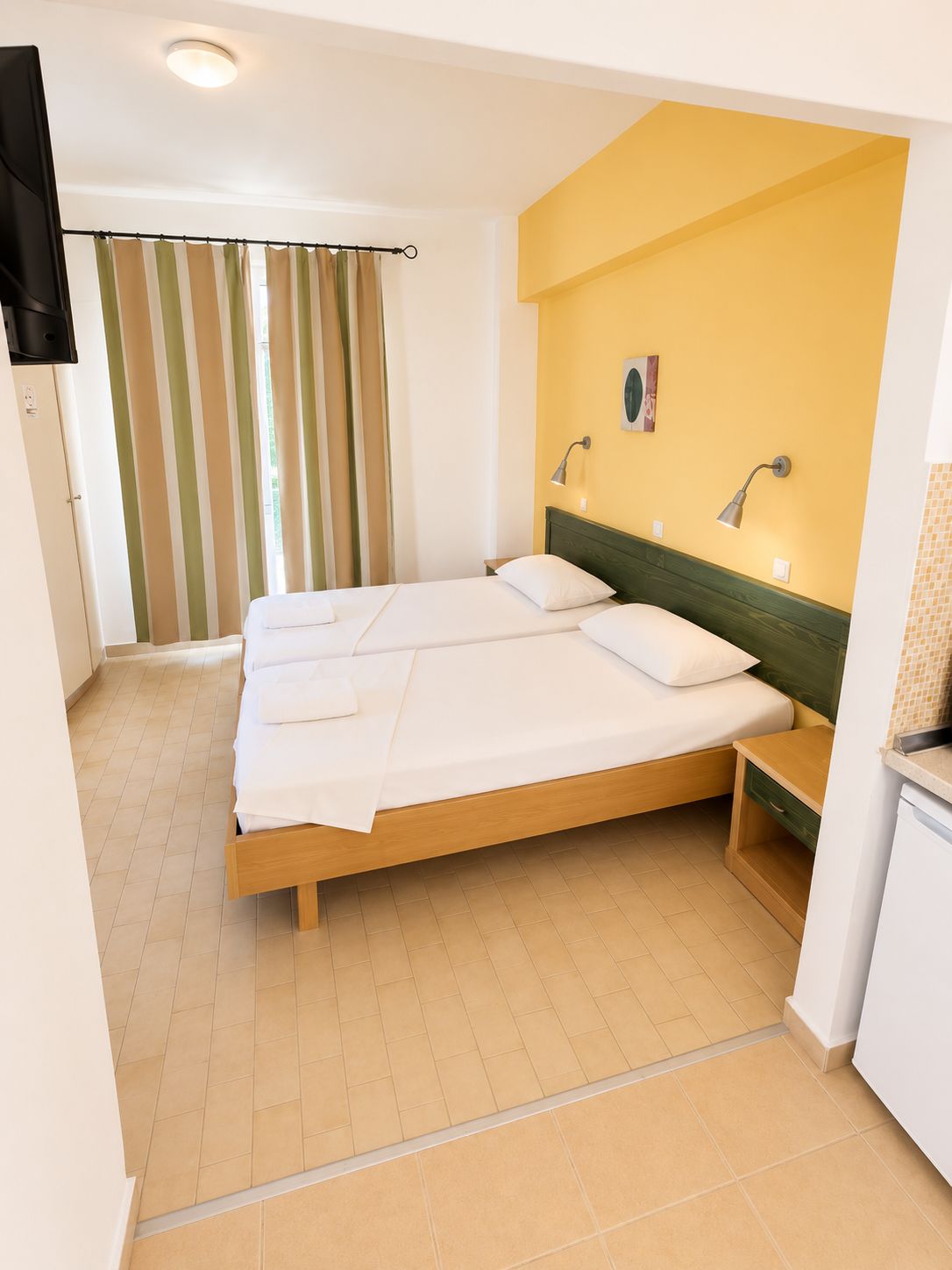 A hotel room with a double bed, a small bedside table, a wall-mounted lamp, and a television.