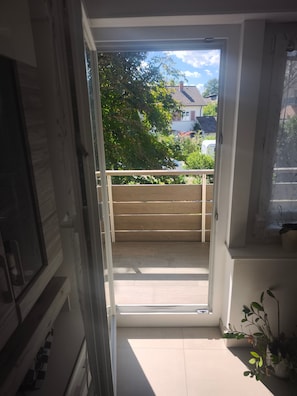 En åben dør, der fører ud til en balkon med gelænder i træ, flisegulv og en potteplante.