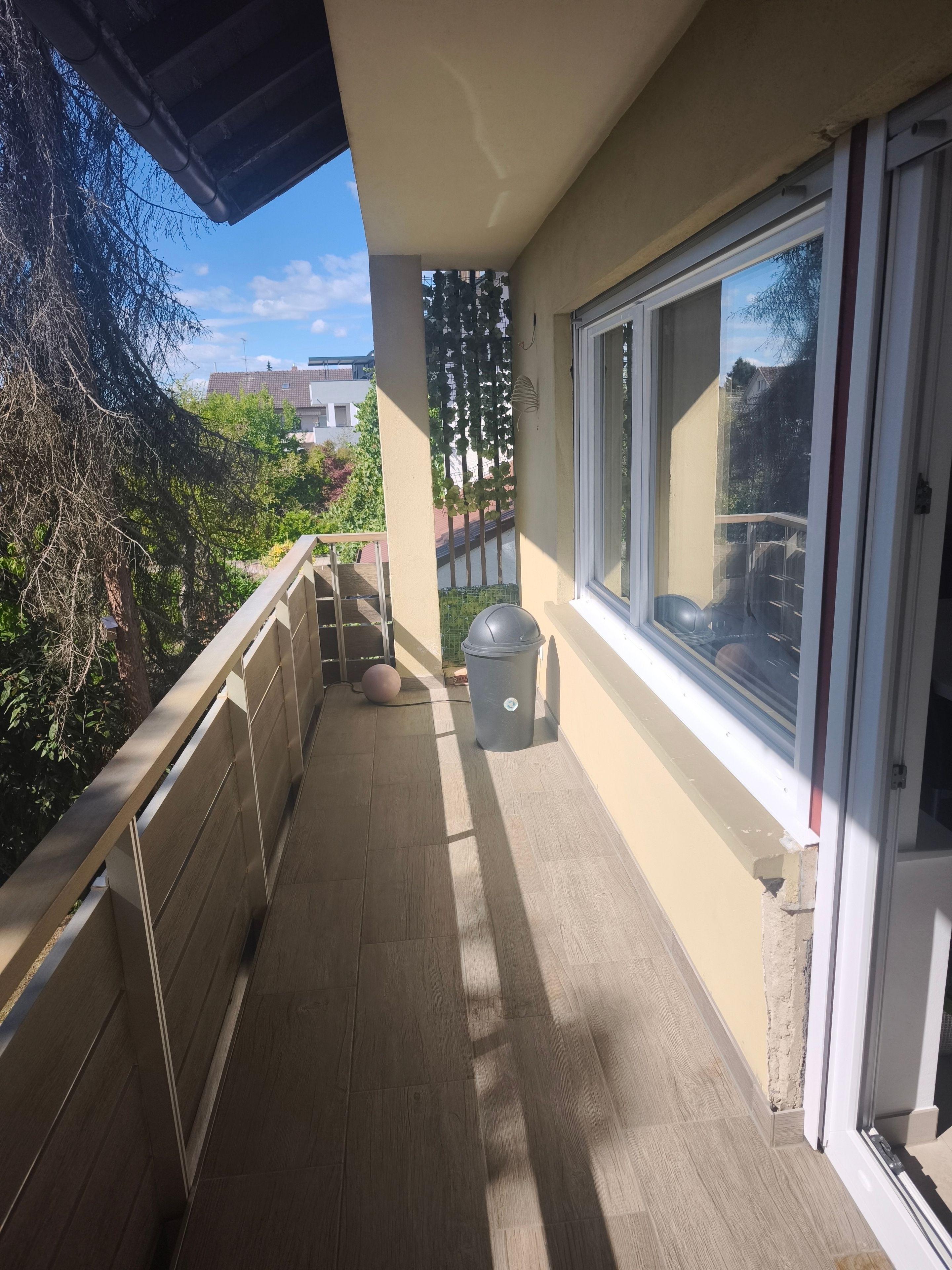 A balcony with a view of the neighborhood, a trash can, and a window.
