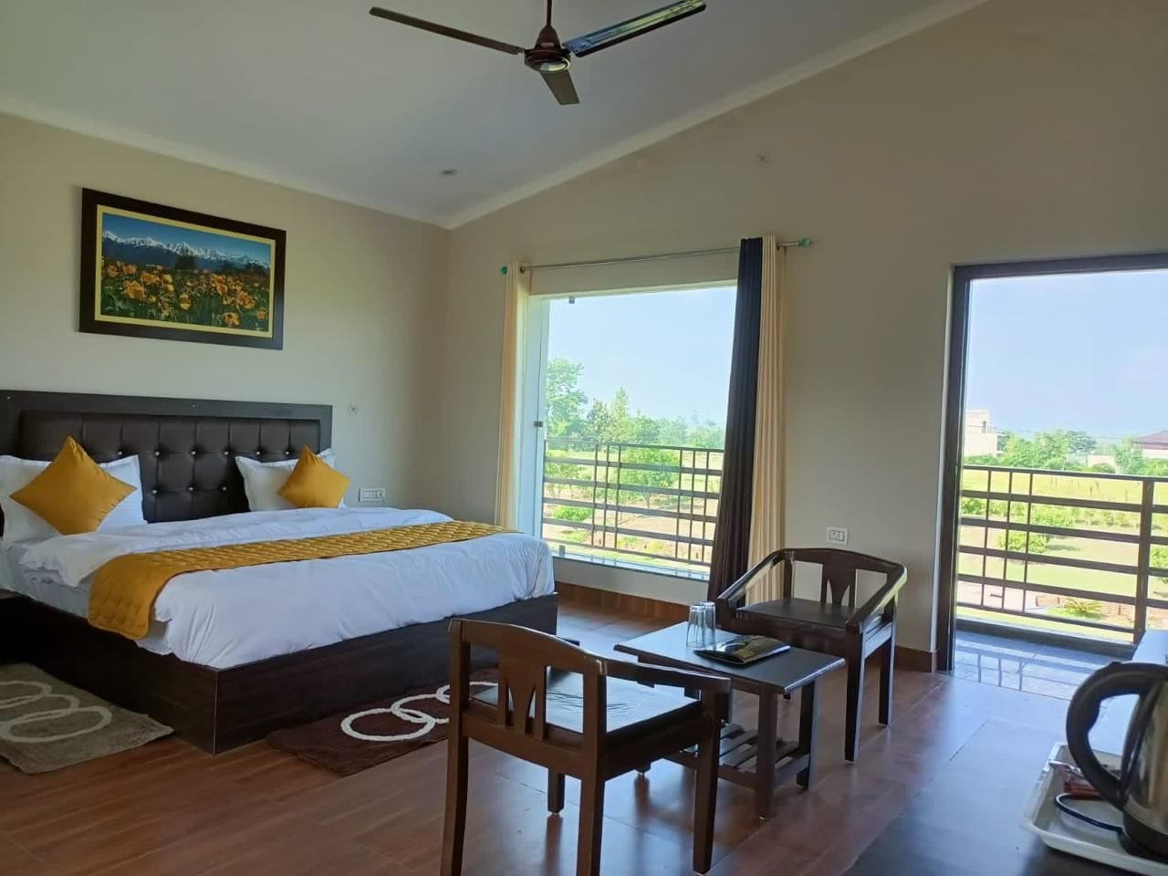 A hotel room with a bed, a desk, chairs, a ceiling fan, and a large window with a view.
