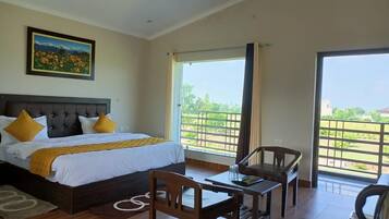 A hotel room with a bed, a desk, chairs, a ceiling fan, and a large window with a view.