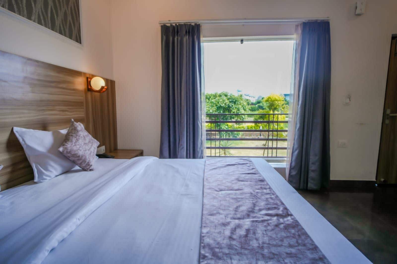 A hotel room with a bed, a nightstand, a lamp, and a view of greenery through the window.