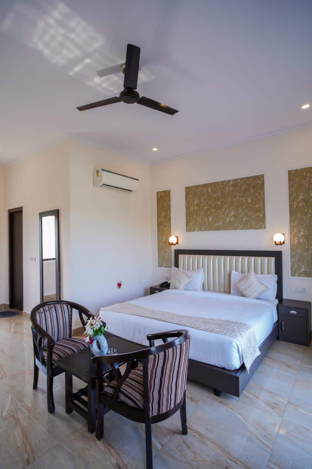 A hotel room with a bed, bedside tables, a desk with chairs, a ceiling fan, and artwork on the walls.
