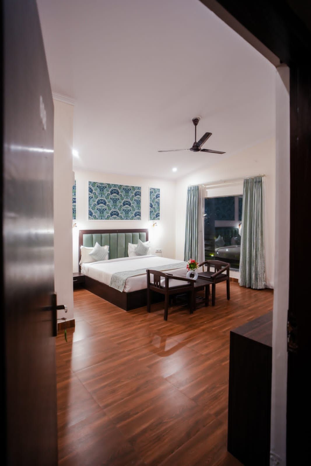 Habitación de hotel con cama, una mesa y sillas, ventilador de techo y ventana con cortinas.