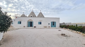 A white building with three conical roofs and blue shutters, a gravel driveway, and a stone wall in the background. - Trulli in Puglia With Private Forest (Locorotondo)