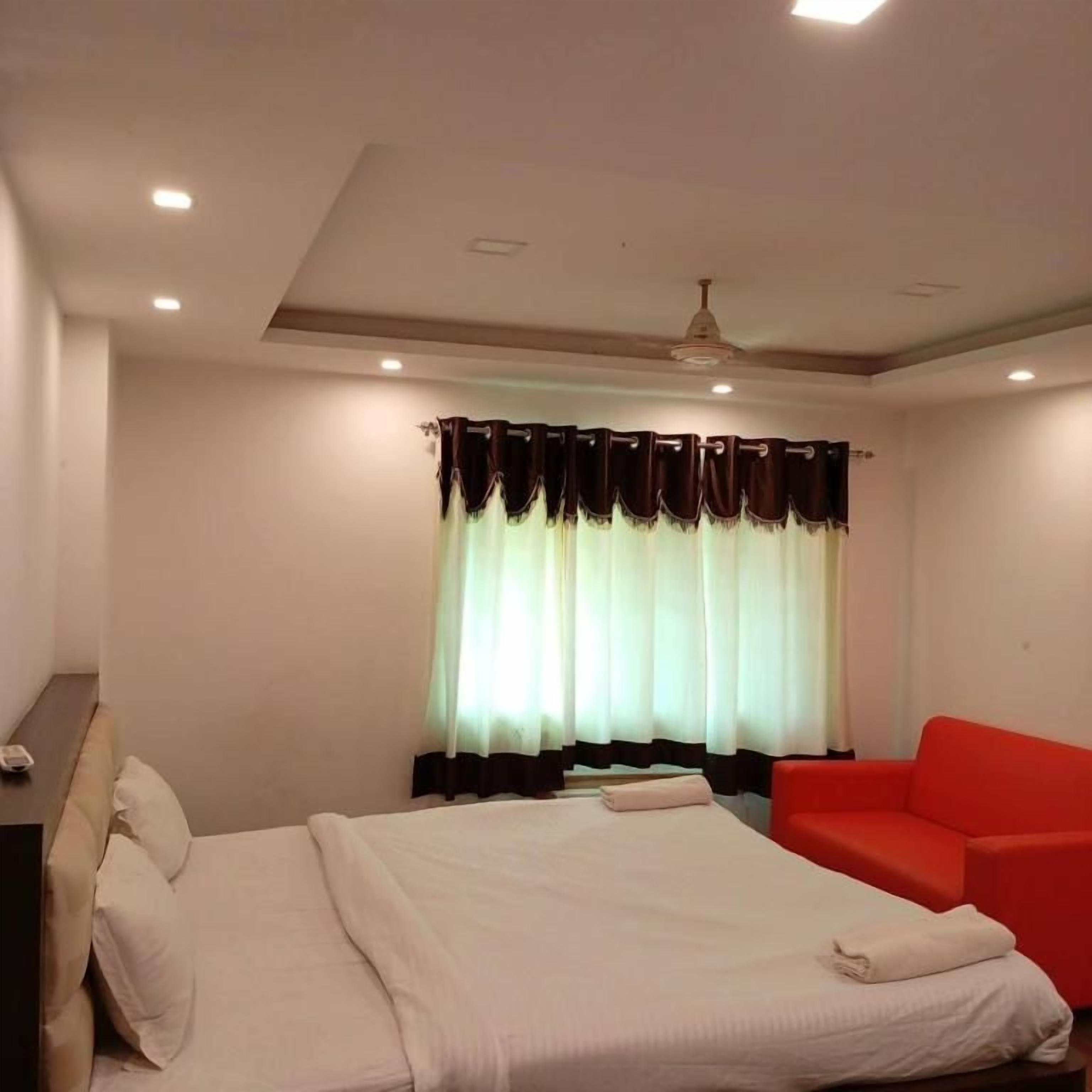 A hotel room with a bed, a red sofa, a window with curtains, and recessed ceiling lights.