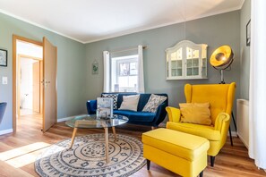 A living room with a blue sofa, a yellow armchair, a glass coffee table, a patterned rug, and a white cabinet with glass doors. - Ferienwohnung in St.peter-ording (St. Peter-Ording)