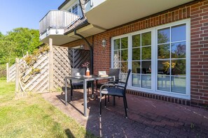 A patio with a table and chairs, a wooden fence, and a brick building with large windows. - Ferienwohnung in St.peter-ording (St. Peter-Ording)