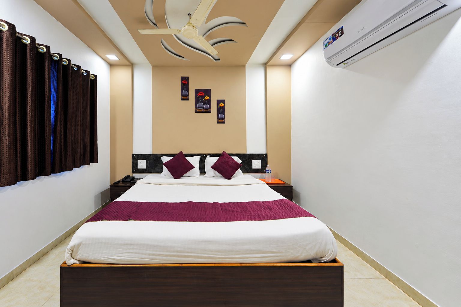 A hotel room with a bed, a nightstand, a wall-mounted air conditioner, and a ceiling fan.