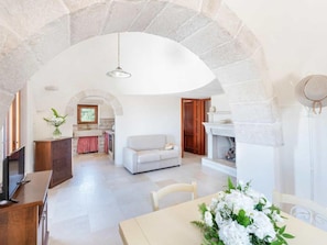 A spacious living area with a stone archway, a fireplace, a dining table with a floral centerpiece, and a television. - Trullo in Itria Valley Near Ostuni Beaches (Ostuni)