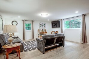 Ein Wohnzimmer mit einer Couch, einer Lampe, einem Teppich, einem Beistelltisch, einer Wanduhr und einem Fenster mit Vorhängen.