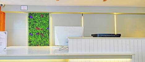 Un comptoir d’accueil avec un plan de travail blanc, une plante décorative fixée au mur avec des fleurs colorées, et un panneau indiquant que l’accès est interdit aux visiteurs.