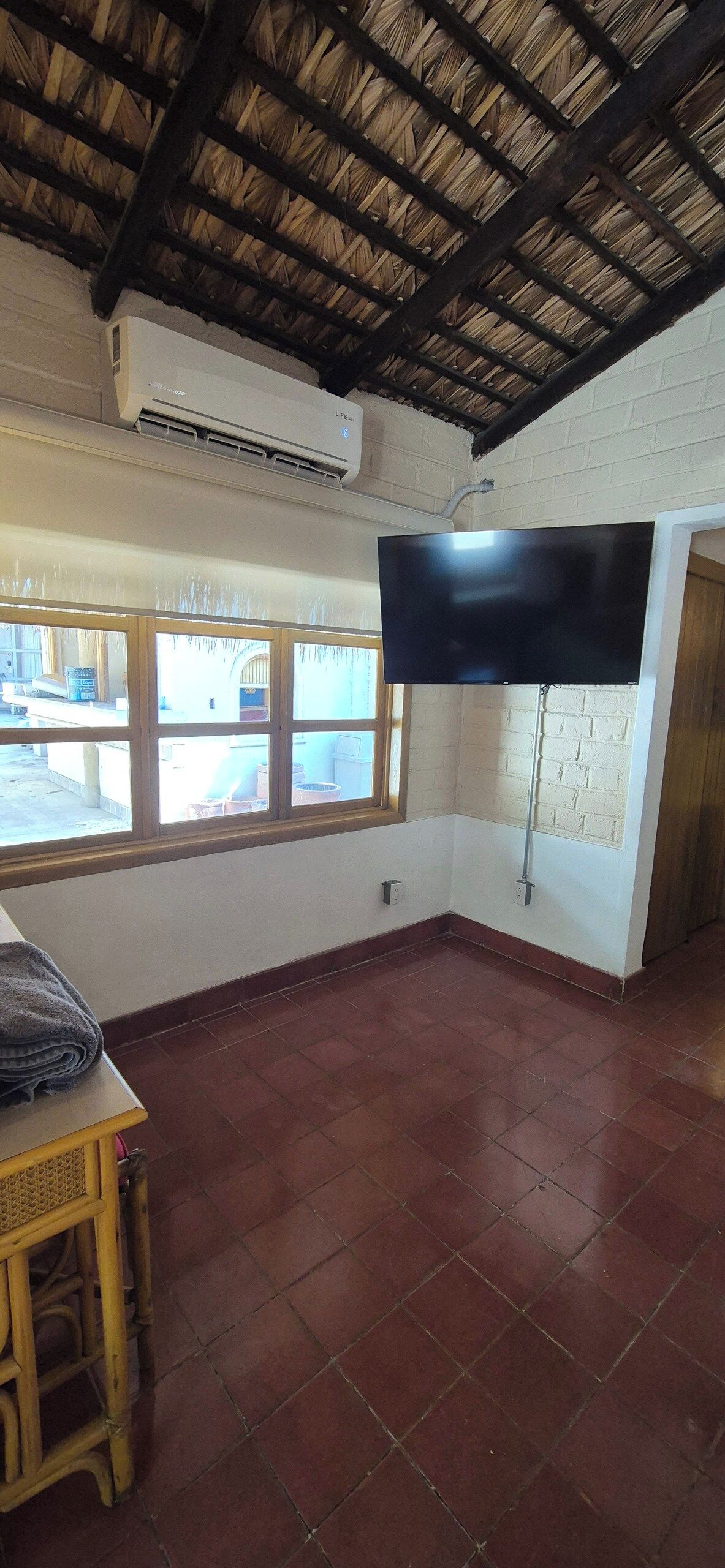 A room with a tiled floor, a flat-screen TV mounted on the wall, a window with a view of the outside, and an air conditioning unit on the ceiling.
