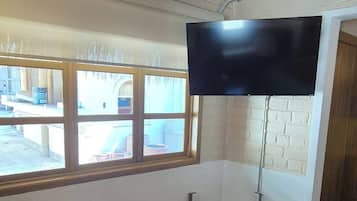 A room with a tiled floor, a flat-screen TV mounted on the wall, a window with a view of the outside, and an air conditioning unit on the ceiling.