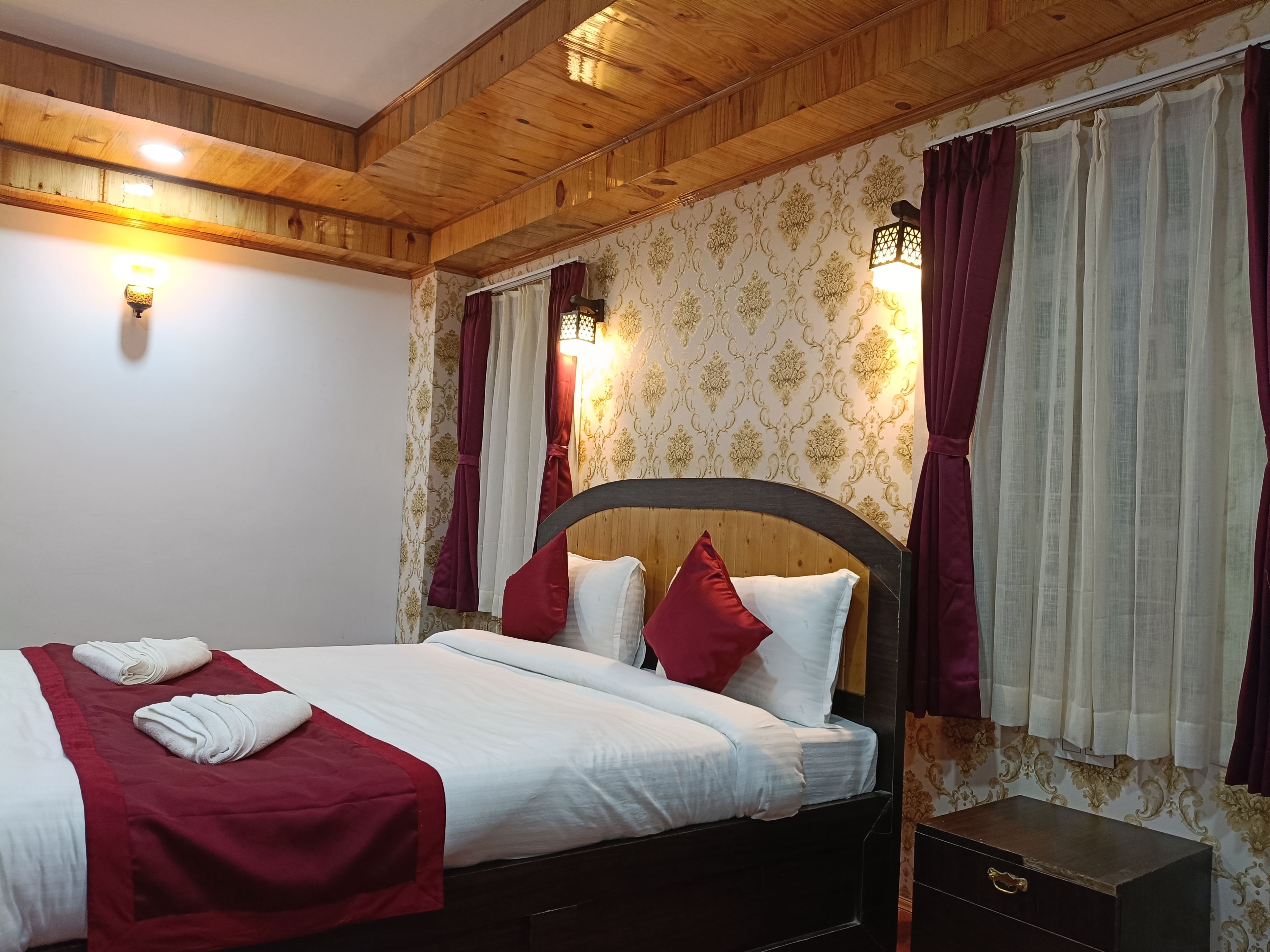 A hotel room with a wooden ceiling, a bed with white bedding and red accents, a nightstand with a drawer, and patterned wallpaper.