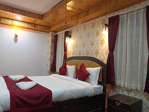 Habitación de hotel con techo de madera, cama con ropa blanca y detalles rojos, mesita de noche con cajón y papel tapiz estampado.