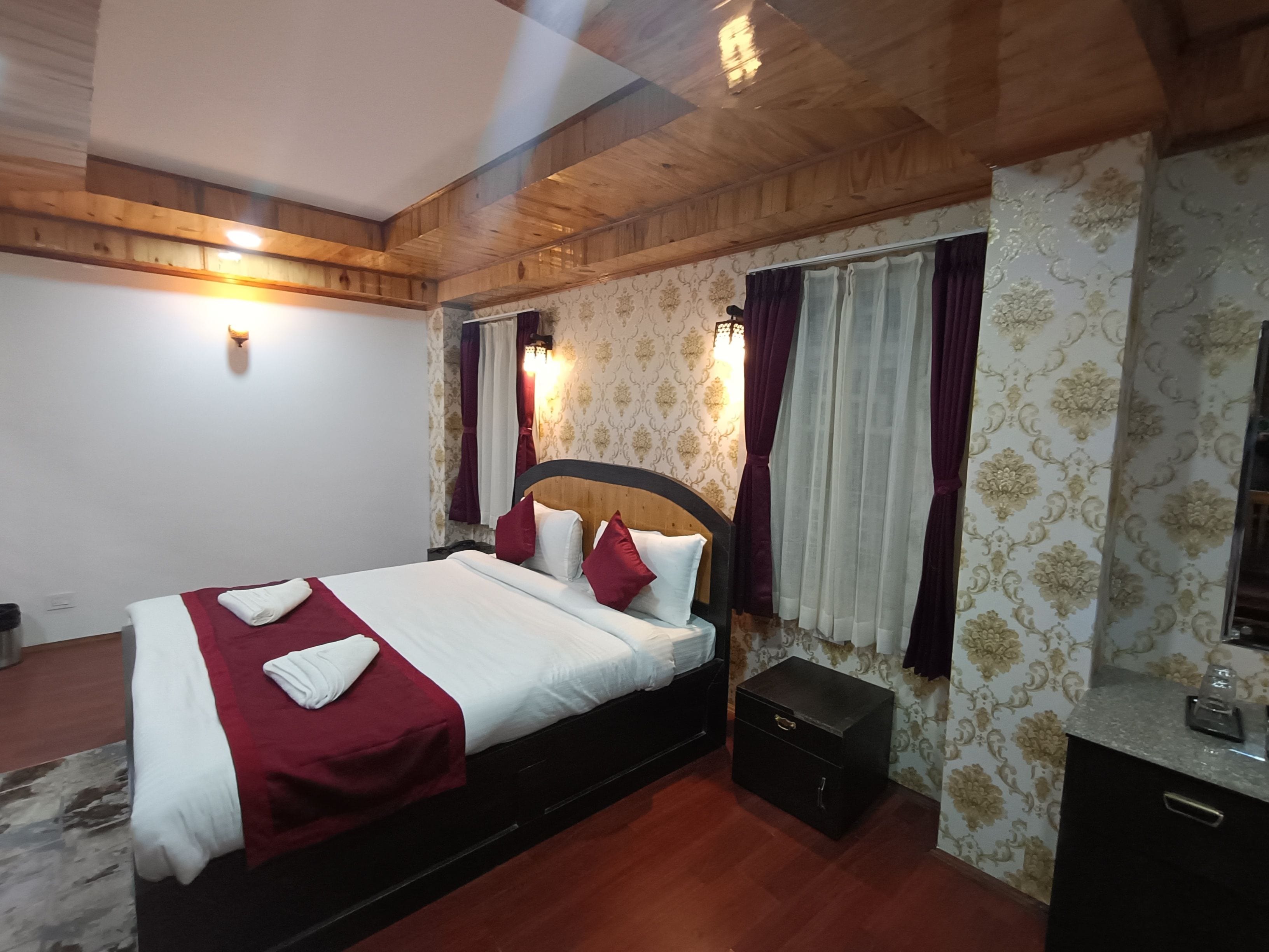A hotel room with a wooden ceiling, a bed with white bedding and red pillows, a nightstand with a drawer, and patterned wallpaper.