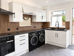 A modern kitchen with a built-in washer and dryer, white cabinetry, and a window providing a view of the outdoors. - Modern 2-Bed House with Garden – Quiet Street, Easy London & Watford Access (Watford)