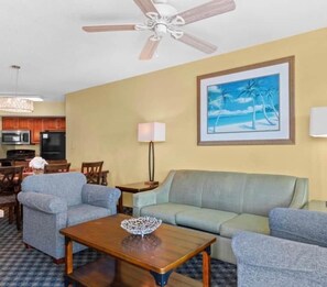 Un salon avec un ventilateur de plafond, un canapé, une table basse et un tableau représentant une scène de plage.