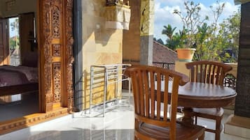 A room with a bed, a wooden chair, a round table, and a balcony with a view of palm trees.