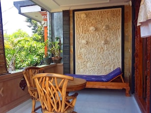 A room with a wooden chair and a bench, a decorative wall panel, and a view of greenery outside. - Puri Asri Bungalow Ubud (Ubud)