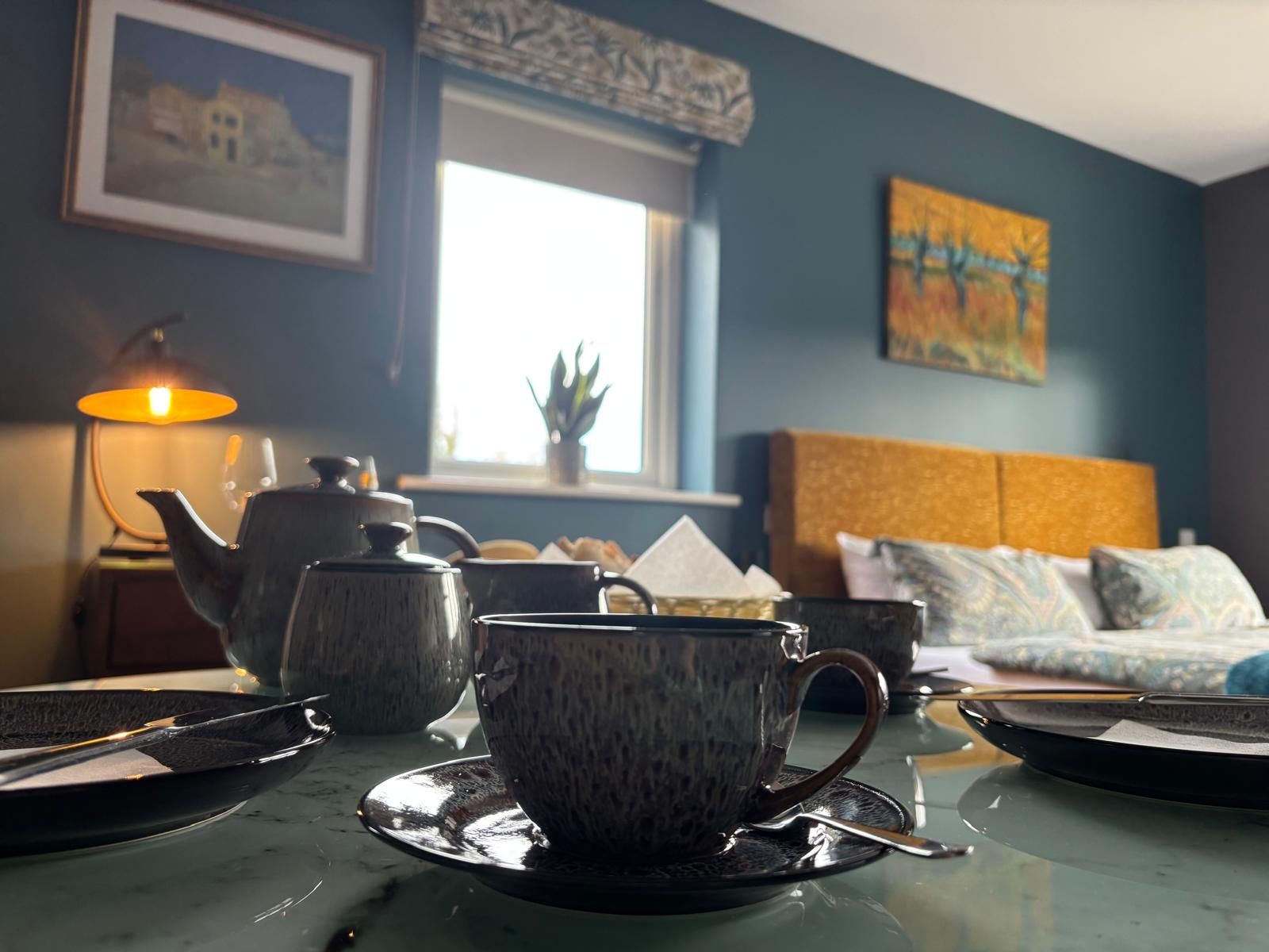 A room with a table set for tea, a sofa, a window, and a painting on the wall.