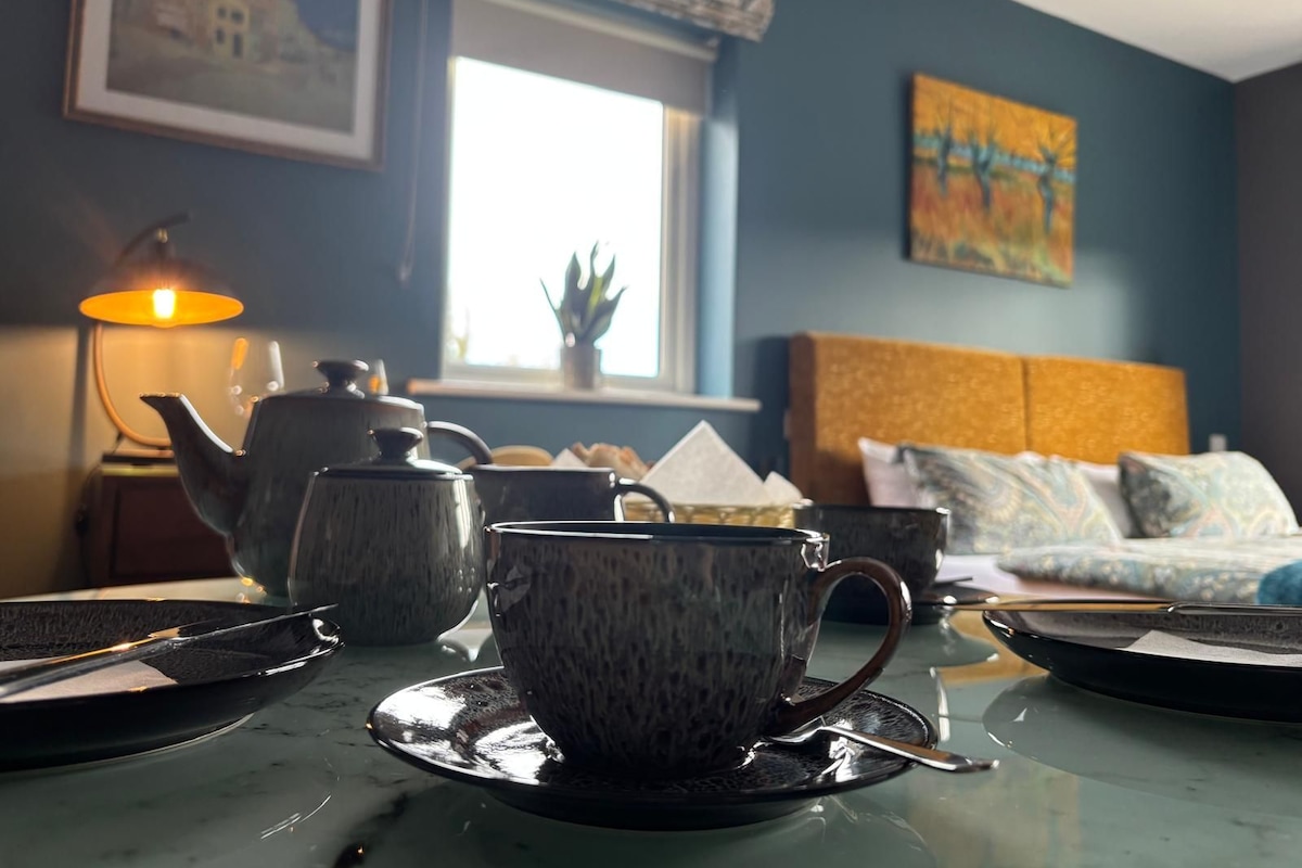 A room with a table set for tea, a sofa, a window, and a painting on the wall.