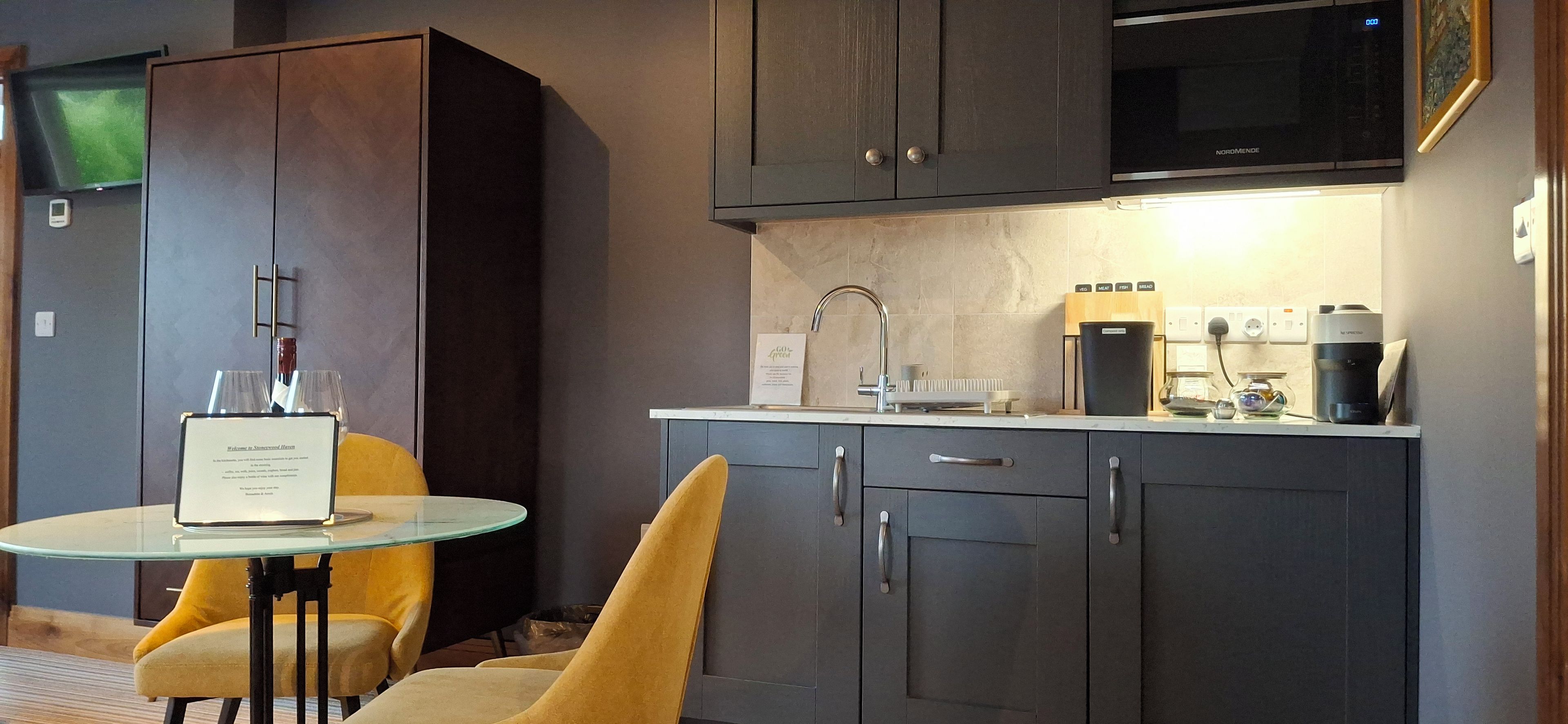Cocina compacta con microondas, cafetera y un pequeño comedor con mesa de vidrio y sillas amarillas.