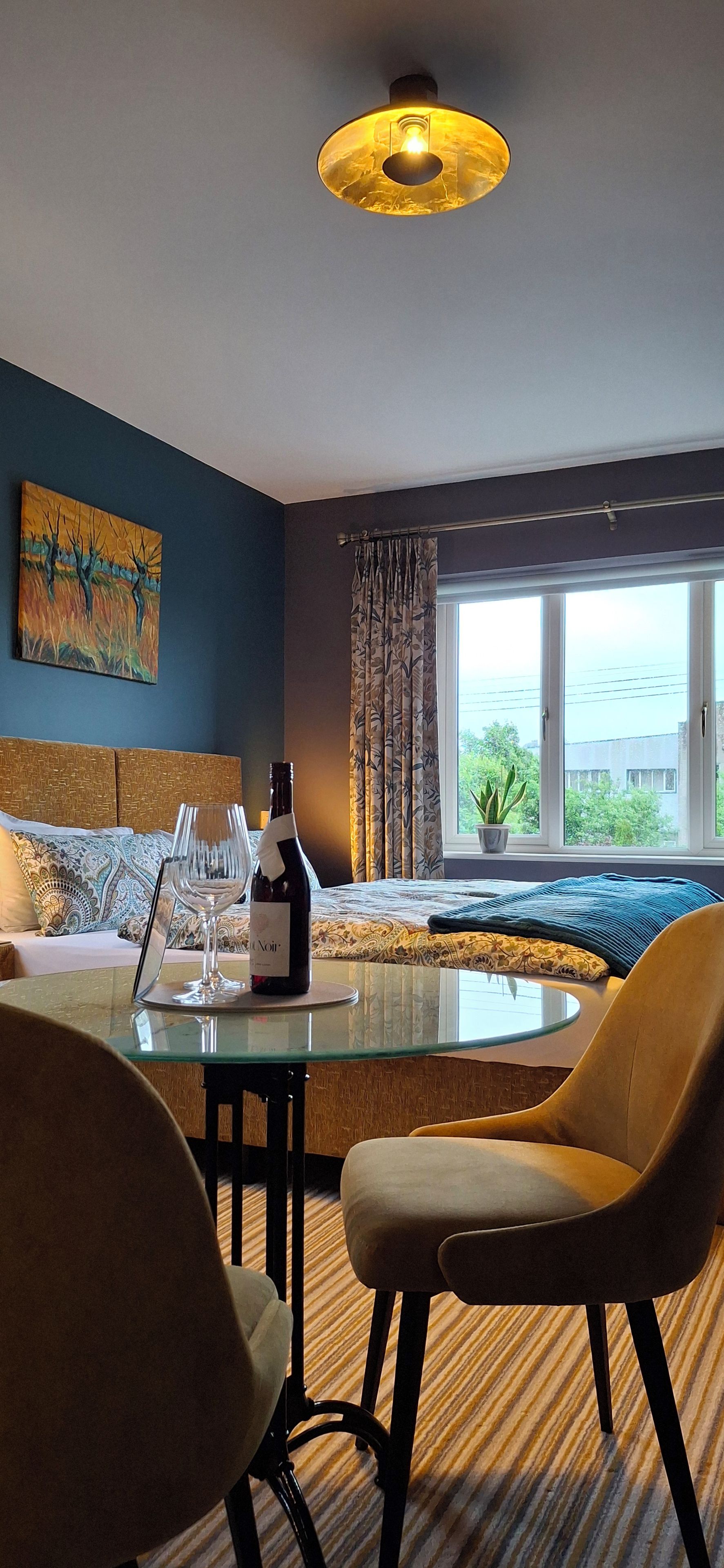 A hotel room with a glass table, chairs, a bottle of wine, a painting on the wall, and a view of the sea through the window.