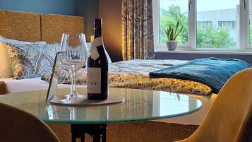 A hotel room with a glass table, chairs, a bottle of wine, a painting on the wall, and a view of the sea through the window.