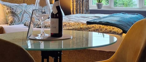 A hotel room with a glass table, chairs, a bottle of wine, a painting on the wall, and a view of the sea through the window.