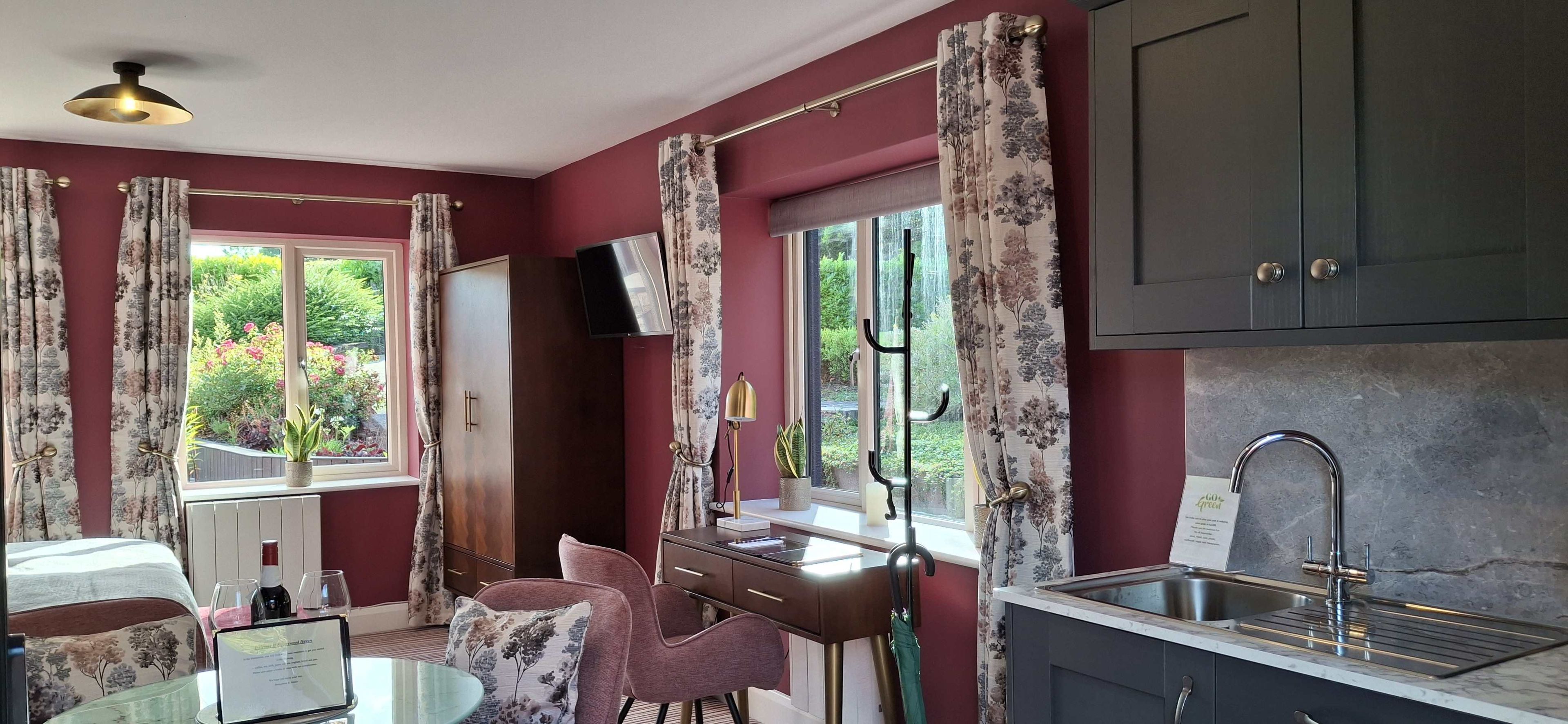 Una habitación con una cama, un escritorio con una silla, una ventana con cortinas, un televisor y una cocina con un fregadero.